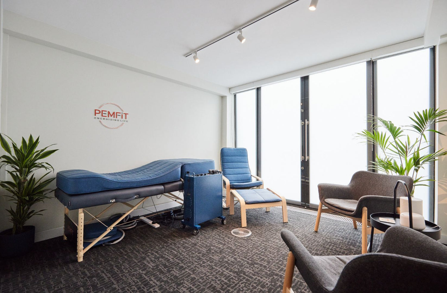 Modern therapy room with blue PEMF bed, chairs, and large windows.